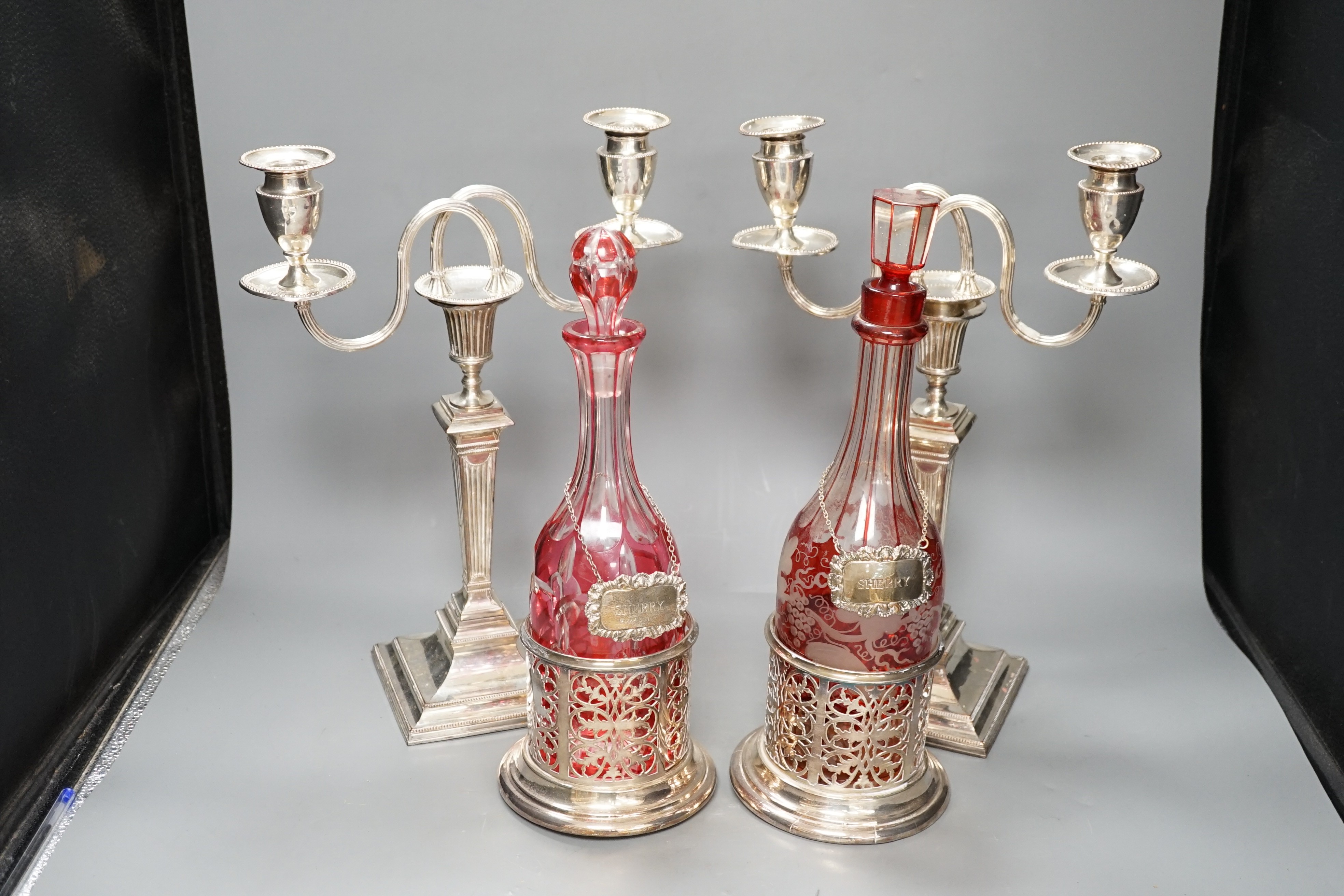 A pair of silver plated two branch, two light candelabra and a pair of silver plated decanter coasters with two ruby flash glass decanters with plated wine labels.